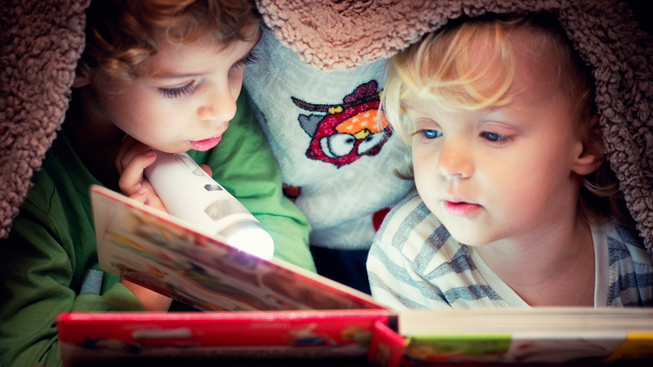 Two children reading a book with a flashlight under a blanket