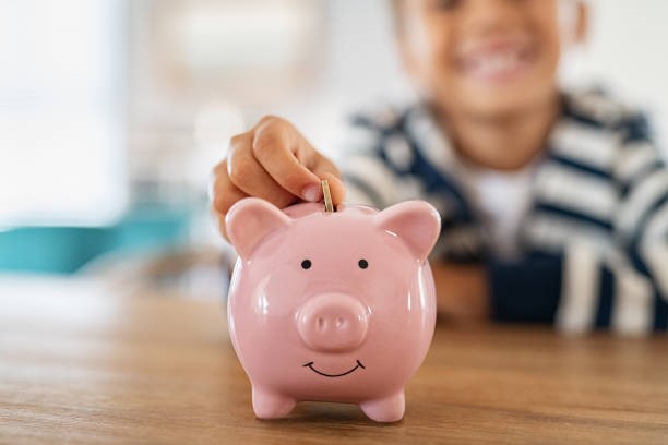 A pink piggy bank in the forefront with a child in the background putting a coin in the bank.