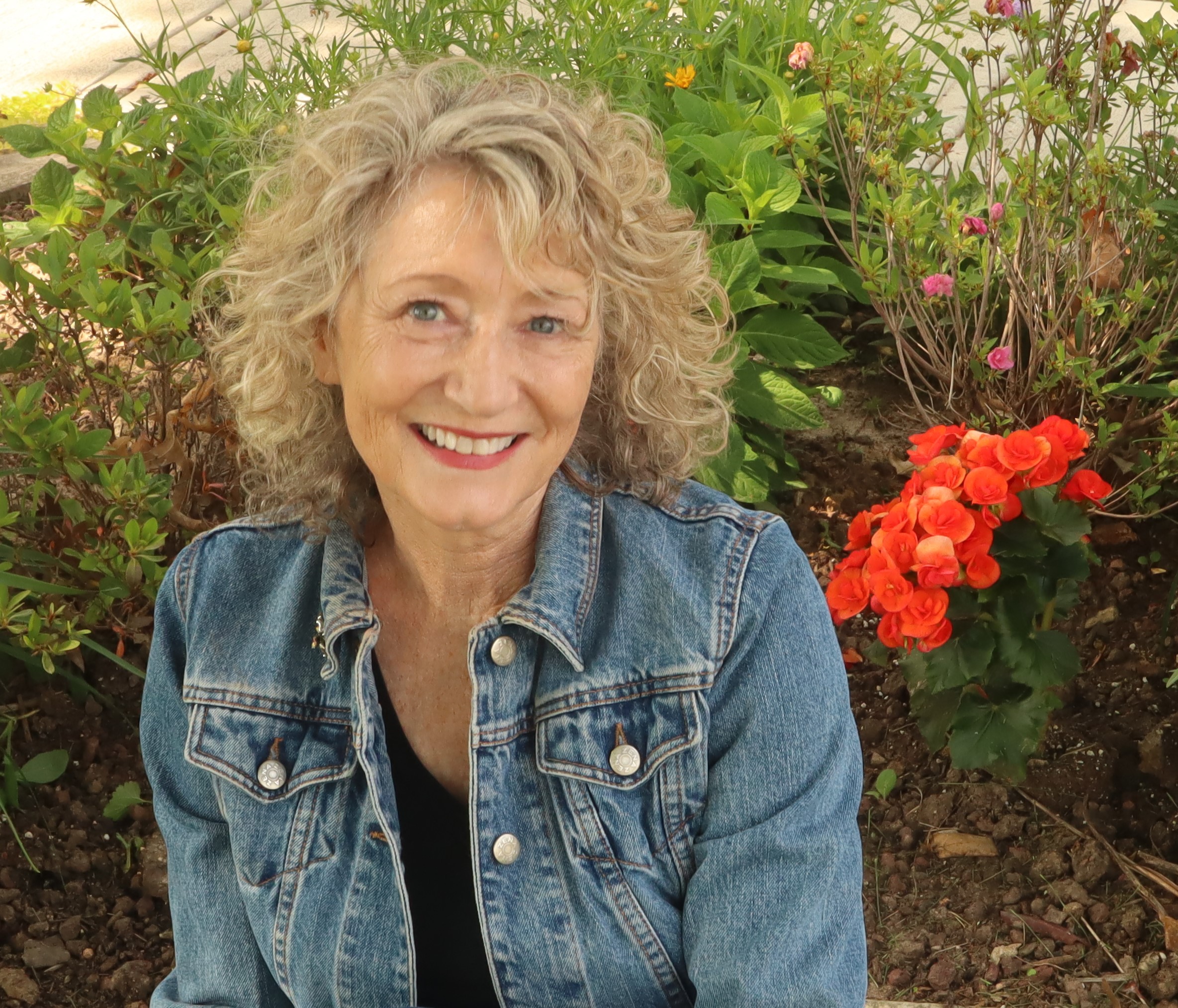 Image of a smiling white woman with short, curly blond hair. She is wearing a black shirt and a jean jacket and sitting in front of bright green and pink foliage. 