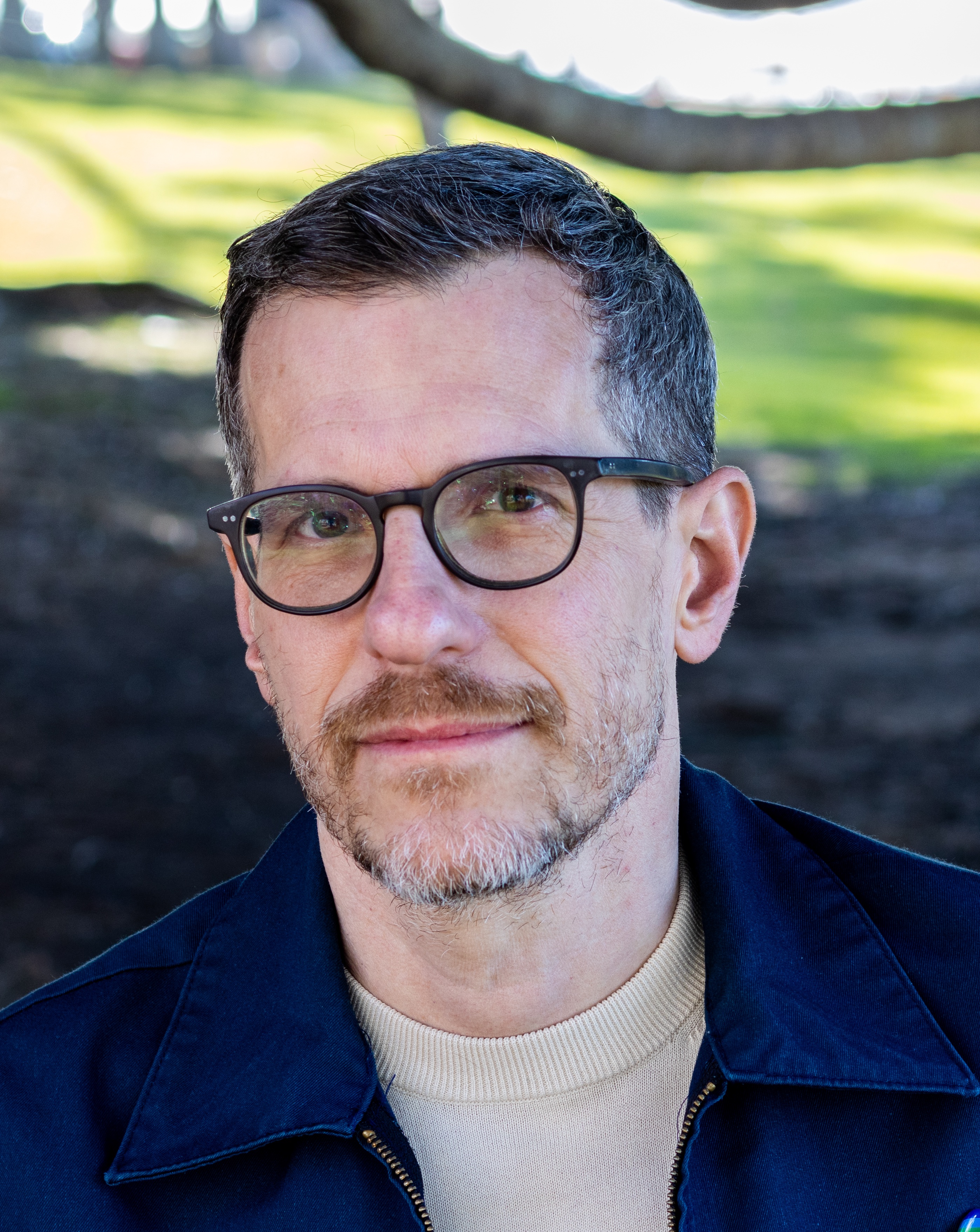 Head shot of a white man with short dark hair, and glasses, wearing an off white shirt with a navy blue jacket. He appears to be standing outside in a park like environment. 