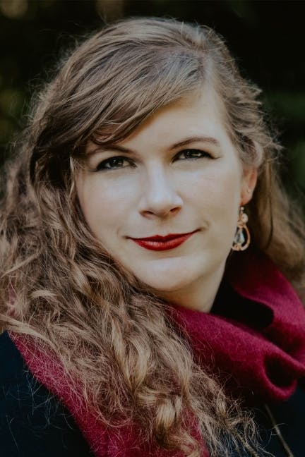 Head shot of a woman with long wavy blond hair. She has on a red or dark pink scarf and a black top. 