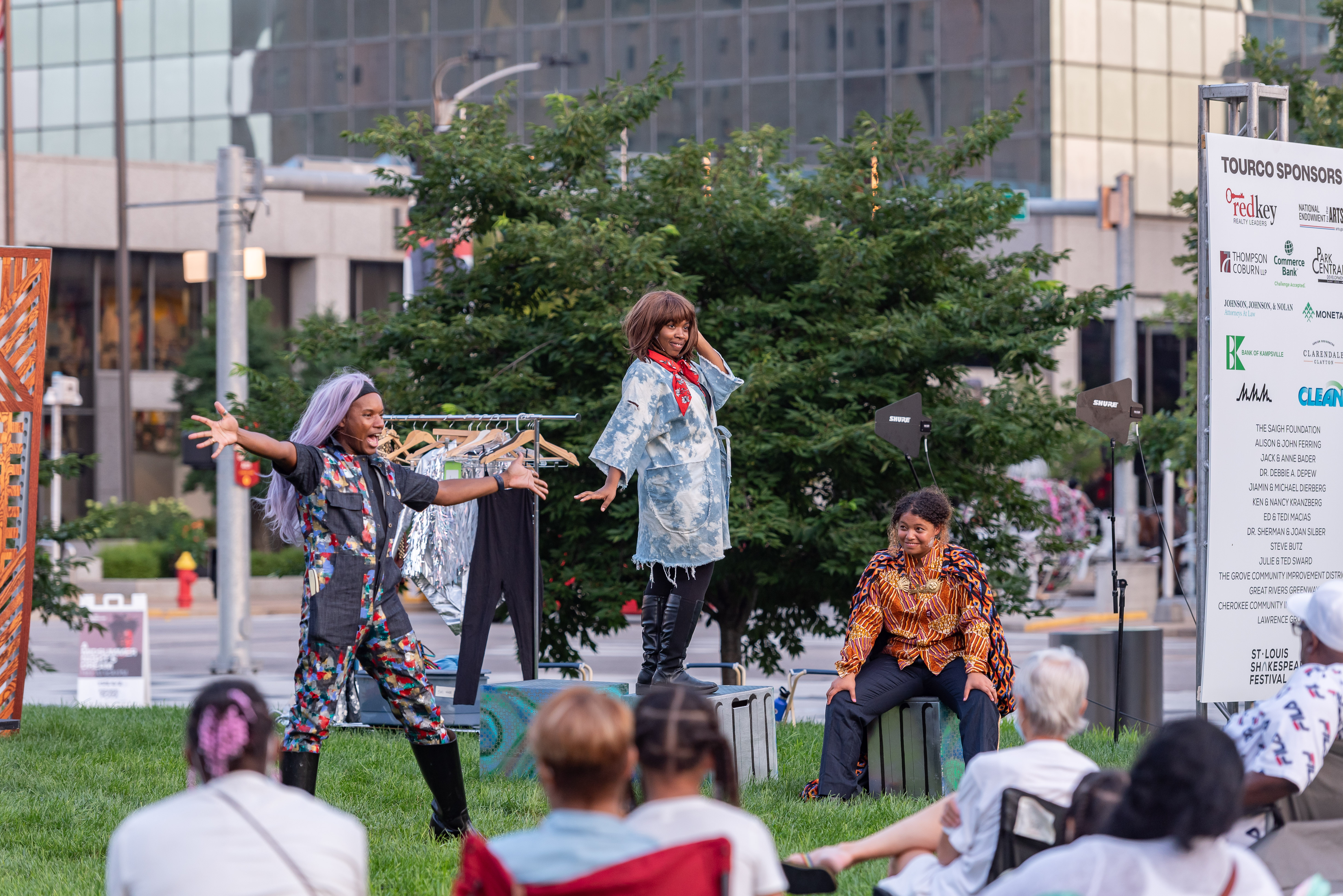 Actors perform outside in a grassy area to an audience seated on the ground and in lawn chairs.