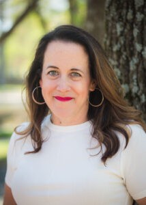 Image of a smiling white woman with long dark hair wearing a white t-shirt standing in front of a tree.