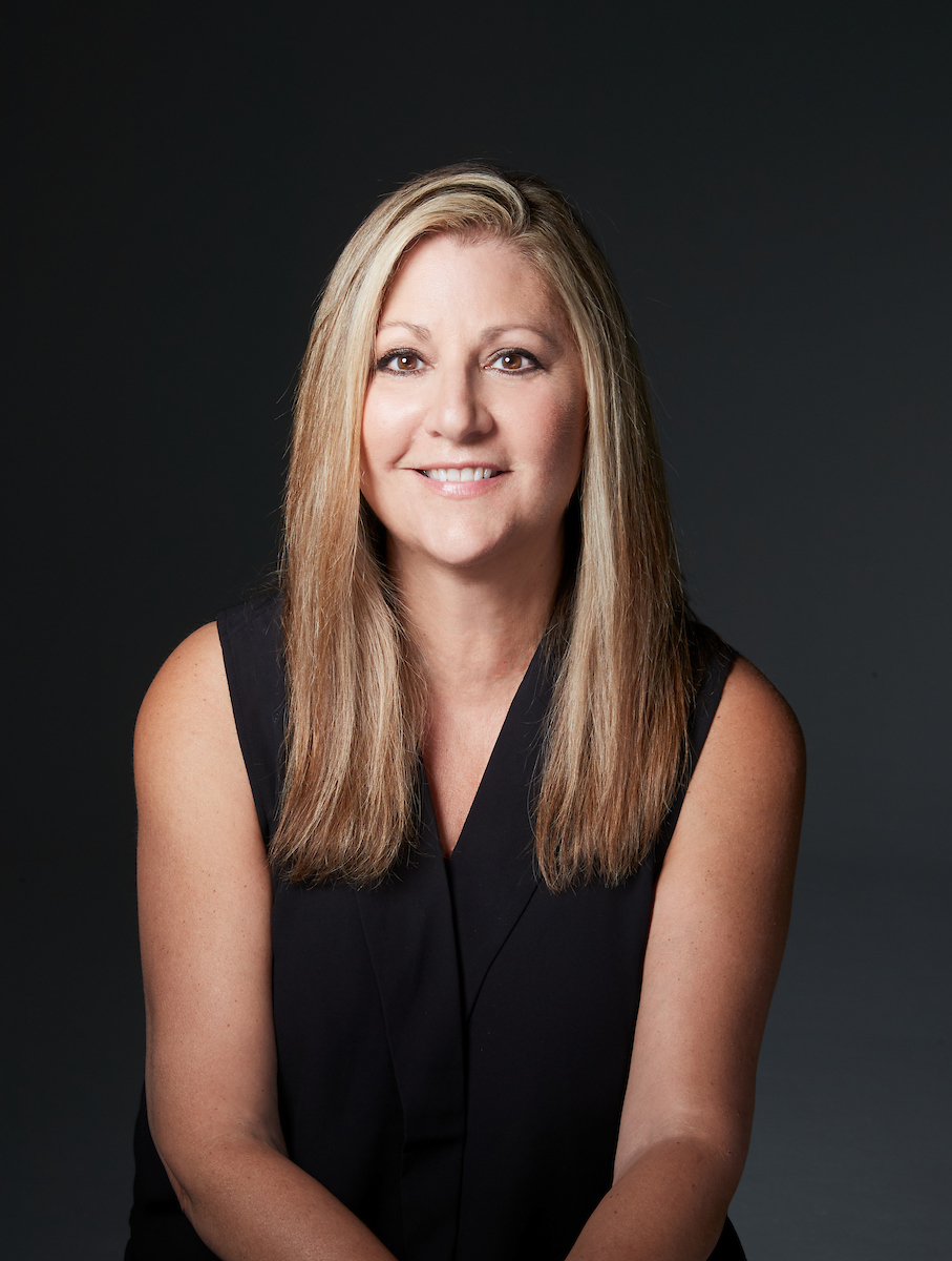 A woman with long blond hair wearing a sleeveless black shirt. She in front of a very dark grey background.