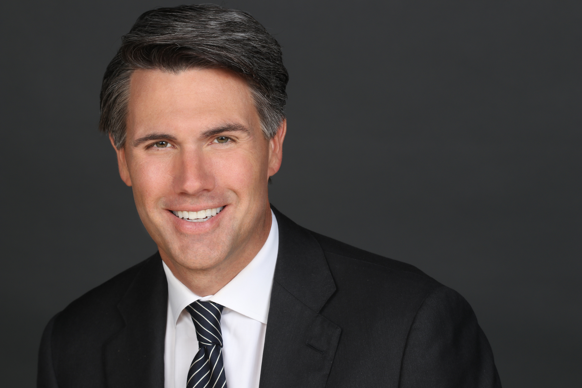 Image of a smiling white man with dark hair wearing a black suit and a striped tie in front of a dark grey background.