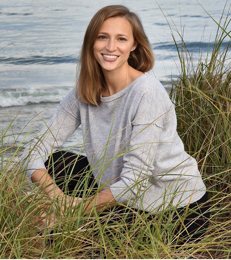 Image of a smiling woman wearing a grey sweater sitting in tall grass with water behind her. 