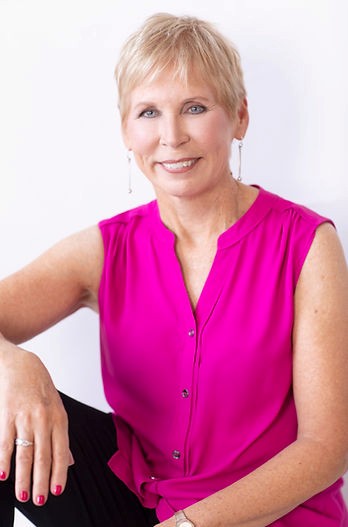 An image of a smiling woman with short blond hair. She is wearing a bright pink sleeveless, button down, tank top and black pants.