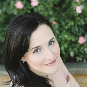 Image of a woman with black hair and blue eyes smiling at the camera. She is wearing a pale pink shirt and standing in front of a pink flowering bush. You can see her head and top of her shoulders. 