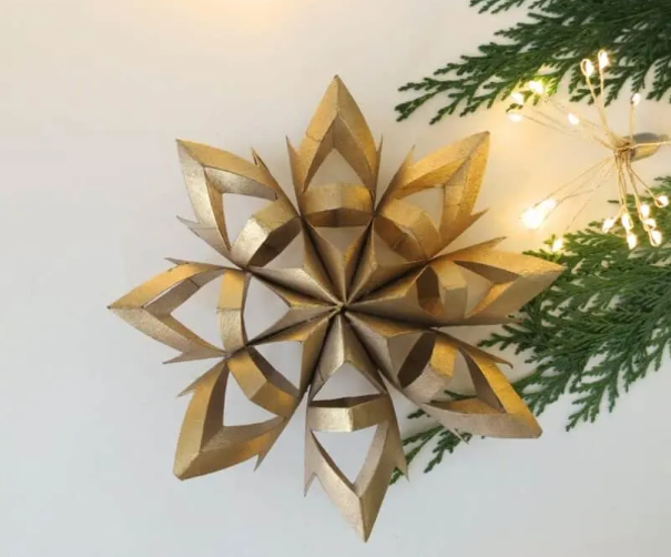 A snowflake decoration made out of toilet paper rolls. There is a warm light on the right hand side of the photo, accompanied by fir tree branches.