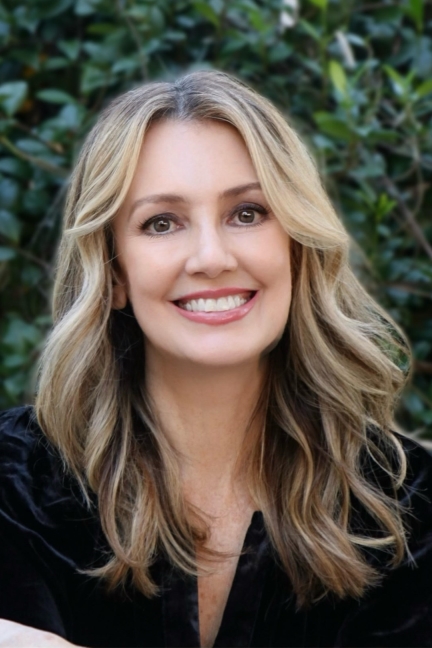Image of a smiling white woman with long blond hair wearing a black shirt. There are leaves in the background. 