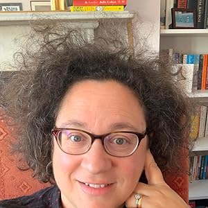 Image of a woman with curly dark hair. She is wearing glasses and sitting in a chair in front of a book shelf. 