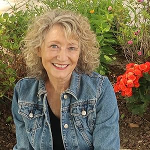 Image of a smiling woman wearing a jean jacket with curly blond hair. Behind her are some red flowers and green bushes. 