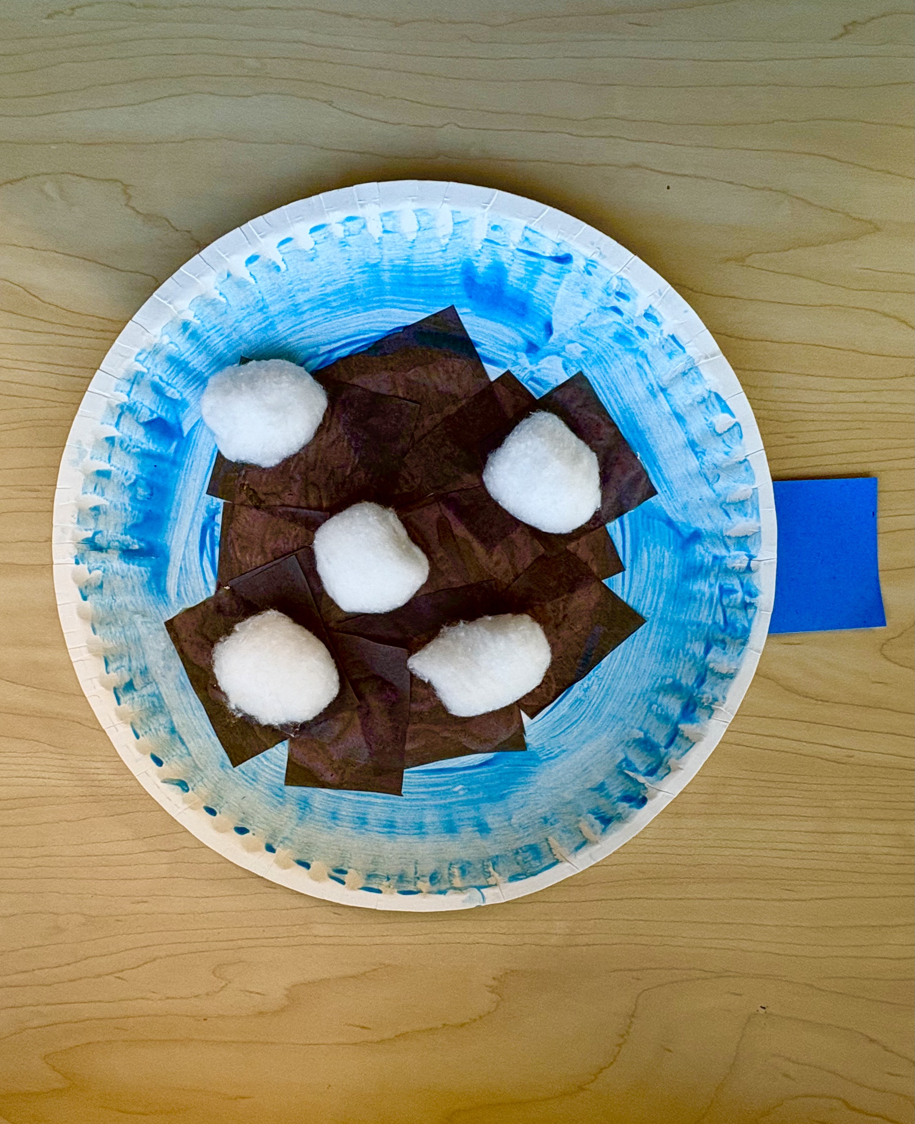 A paper plate painted blue with brown tissue paper and cotton balls on top that resembles hot cocoa