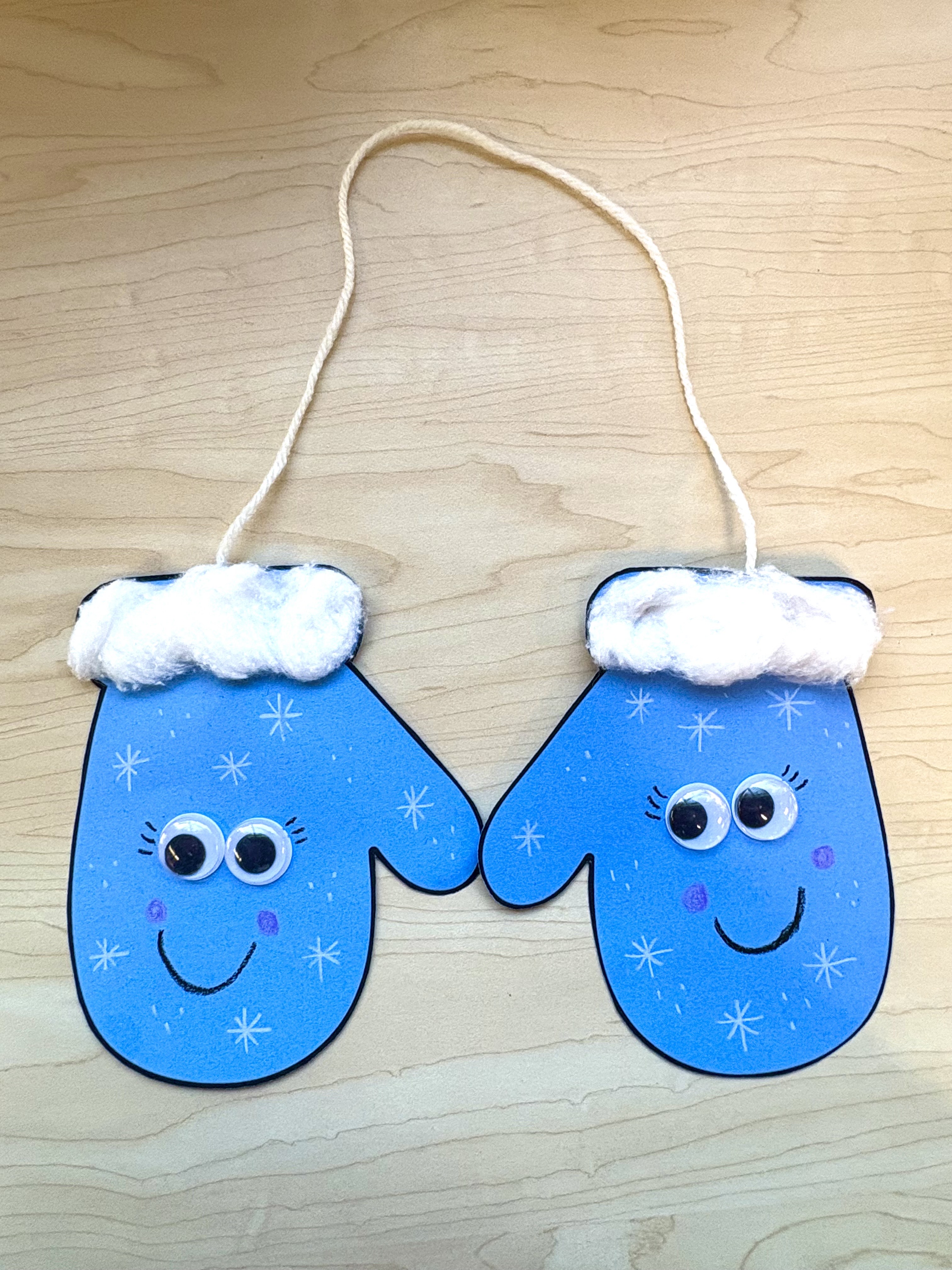 A pair of blue construction paper mittens with googly eyes held together by white yarn