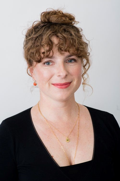 Image of a smiling white woman with brown, curly hair in a bun on the top of her hair. She is wearing a black top and simple gold jewelry.