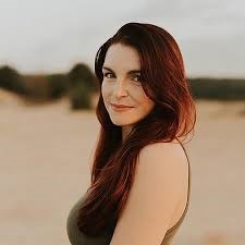 Image a woman with long red hair standing on what looks to be a sandy area.