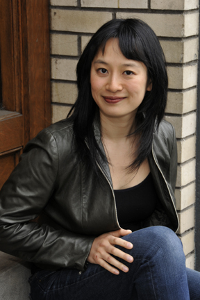 Image of a woman sitting next to a brick wall. She has dark hair and is wearing a black shirt, black leather jacket and blue jeans. 
