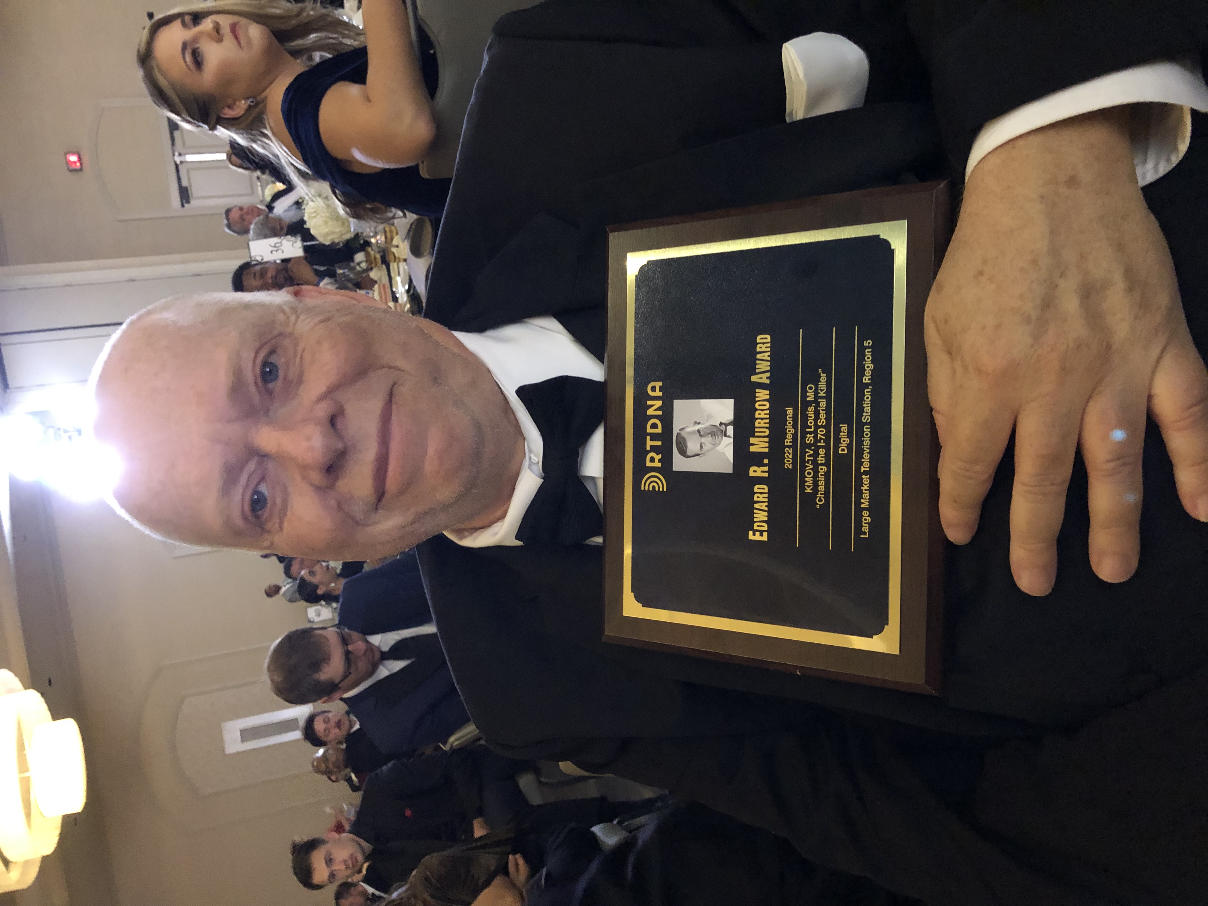 A smiling man in a tuxedo holds an award in a room full of people.