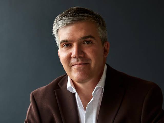 A head shot of a smiling white man in a brown suit and white shirt.