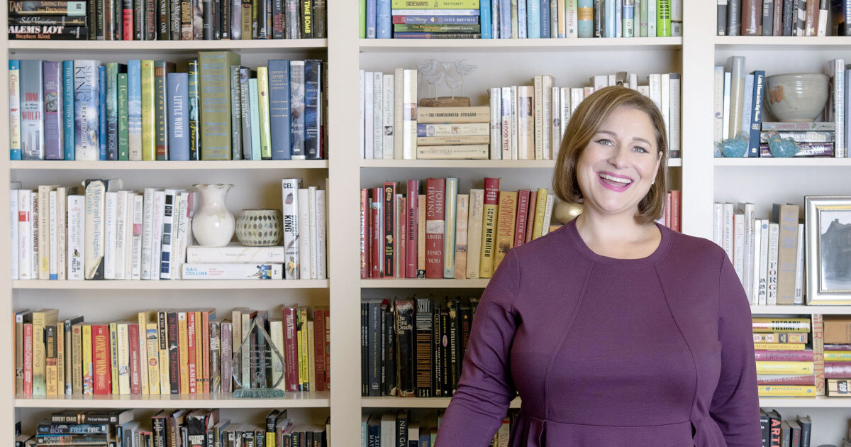 A smiling woman in a purple shirt stands in front of a bookshelf full of books and other items.