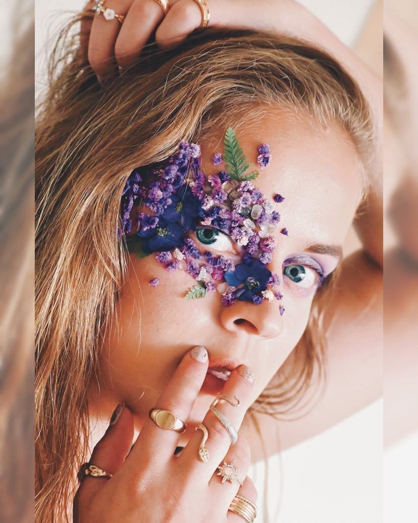 Close up image of a blond woman with purple eye makeup and purple flowers around her eye. She has rings on her fingers and one hand is in her hair while the other hand is near her mouth. 