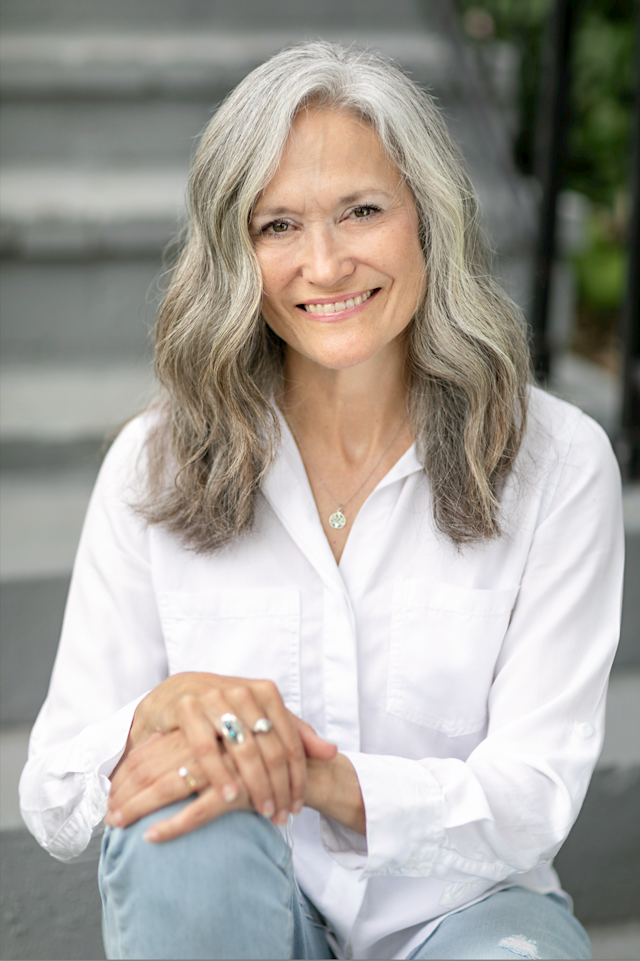 A woman with long grey hair, wearing blue jeans and a white shirts, sits outside on concrete steps. 