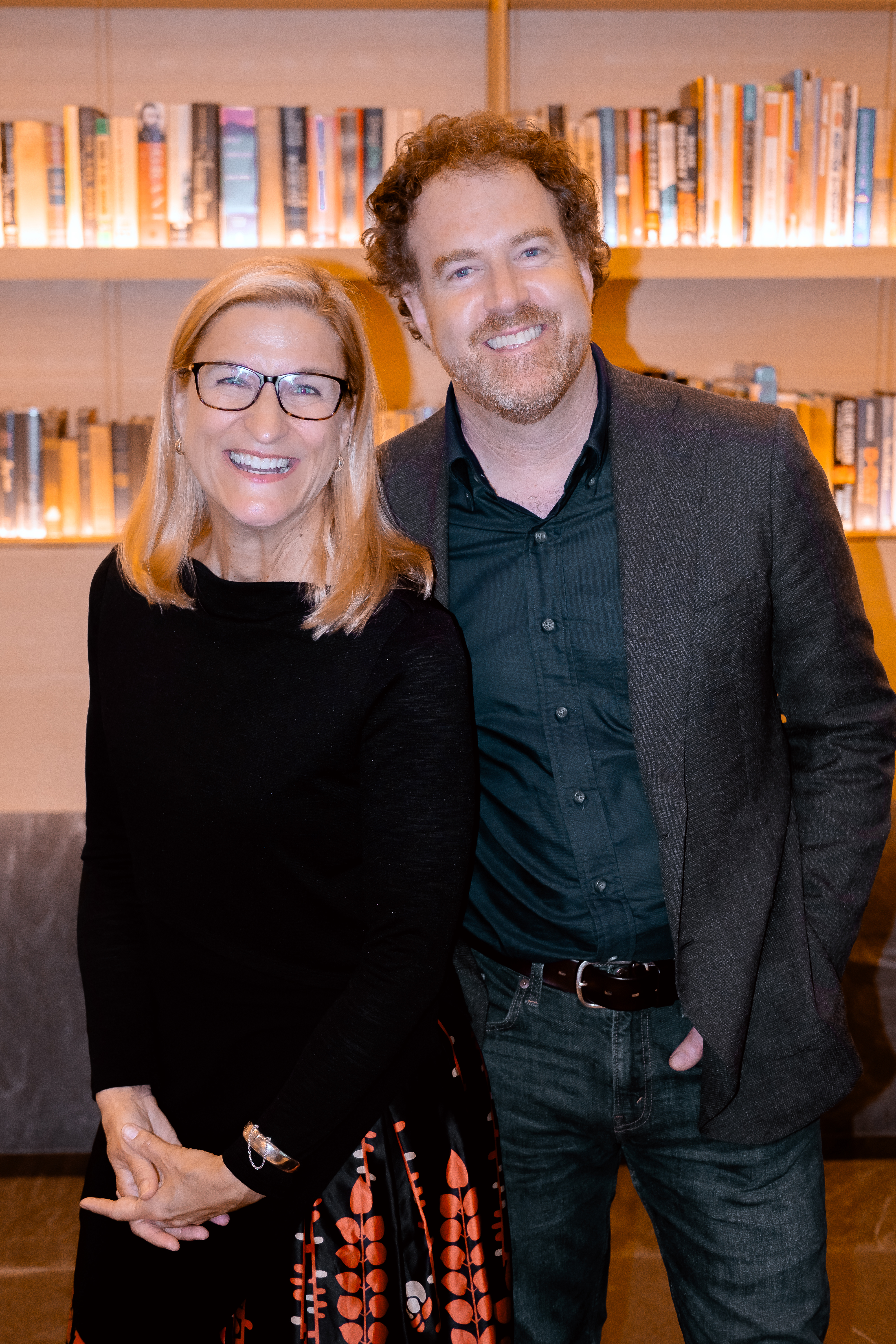 A man with brown curly hair and a woman with blond hair and glasses stand in front of book shelves. They are both smiling at the camera. 