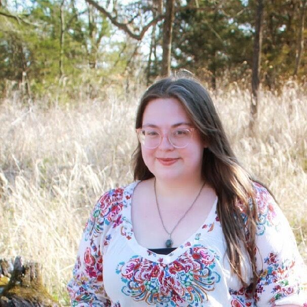 A woman with long brown hair and glasses stands in a field of tall grass and trees. 