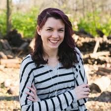 Image of a smiling woman with long dark hair wearing a grey and black striped shirt. She is standing in front of a natural setting with trees.