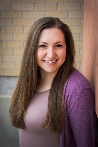 Headshot of a woman with long brown hair wearing a purple shirt standing in front of a brick wall. 