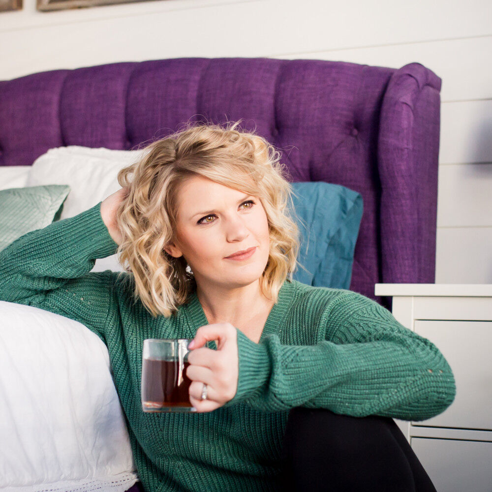 Image of a blond haired woman sitting in front of a purple upholstered bed with white blankets and blue pillows. She is wearing a green shirt and black pants and is holding a mug with a dark colored drink in it. 