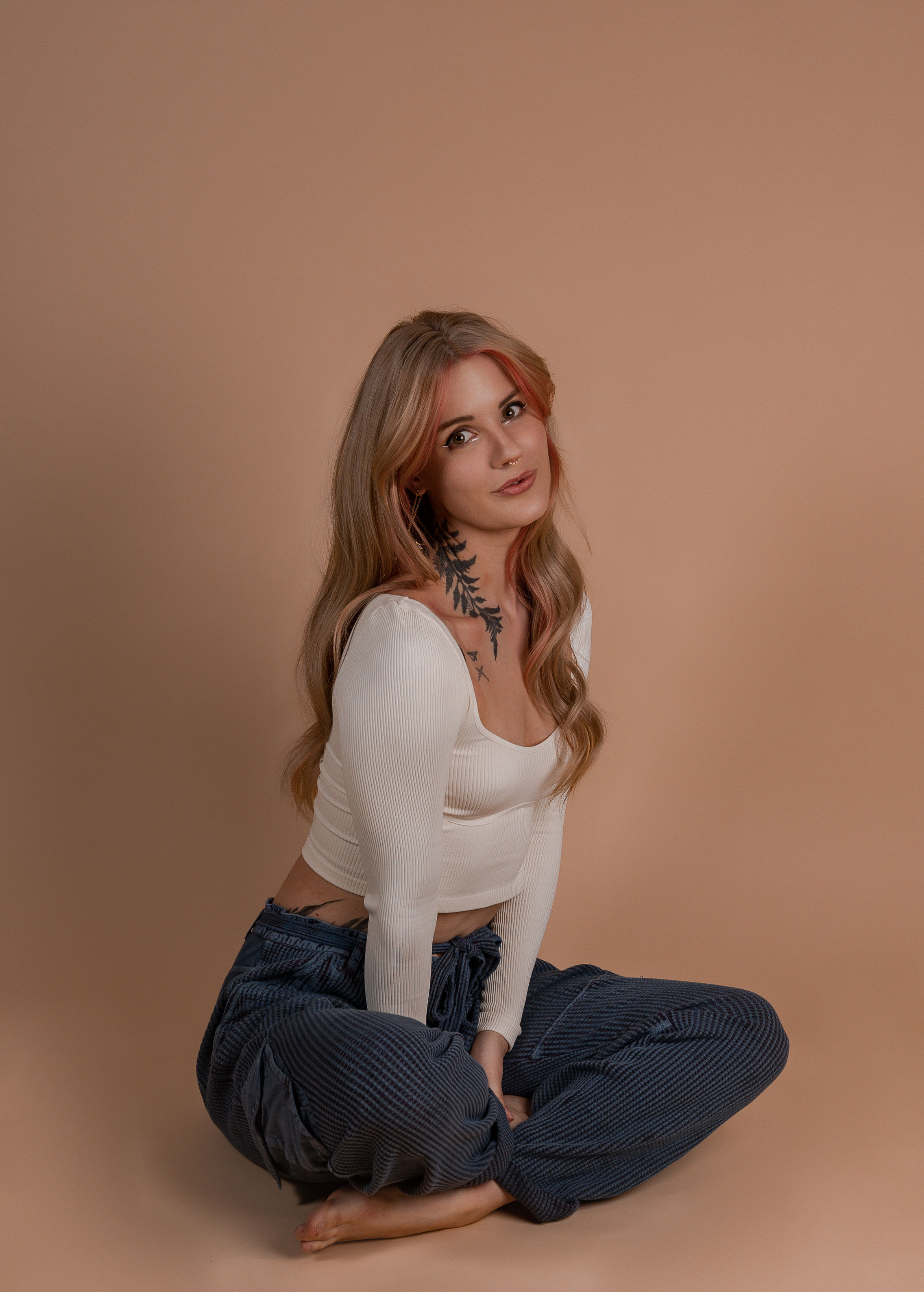 Image of a woman sitting cross legged on the floor. She has long blonde hair with pink highlights and is wearing a white shirt and blue corduroy pants. She has a large tattoo of a branch with leaves on her neck. 