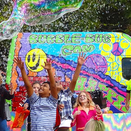 Kids reach for bubbles in front of a colorful bus. 