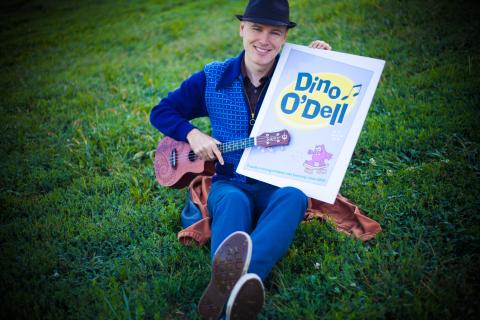 A man in a hat sits in a field with a ukulele and a sign. 