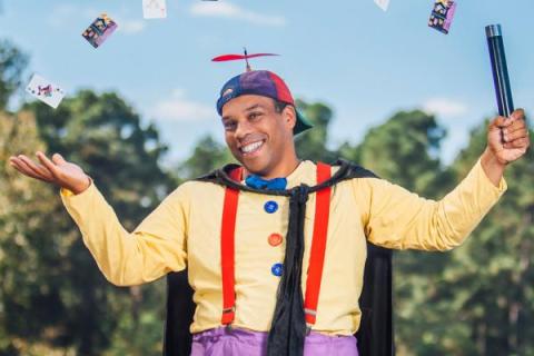 An African American man juggling in a yellow shirt with suspenders, a cape, and a funny hat. 