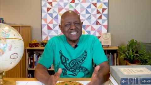 Image of a smiling black man in a teal shirt. Behind him, hanging on a wall is a quilt. On one side of him sits a globe, on the other side is a stack of books. 