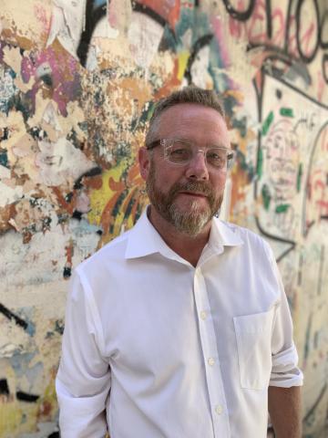 Image of a man wearing a white button down shirt. He has on glasses and has a beard. His hair is light brown. He is standing in front of a colorful, abstract painted wall. 